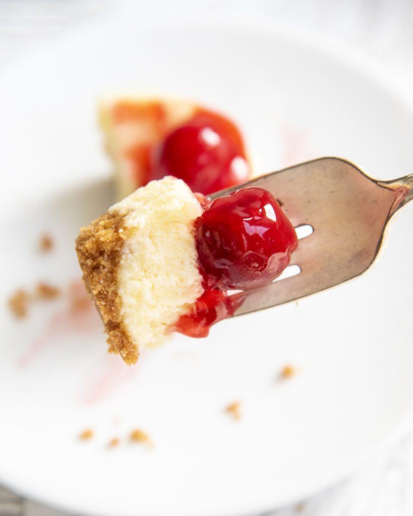 A bite of cheesecake on a fork topped with a pie filling cherry.