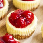 A close up of mini cherry cheesecakes, with ridges from a muffin paper liner on the sides, and 4 canned cherries on top.