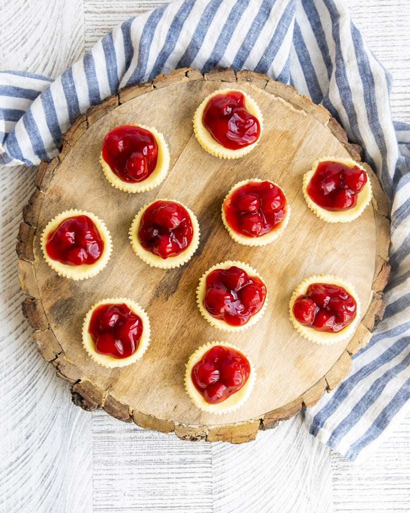 An overhead photo of 10 mini cherry cheesecakes on a round wooden board, showing the edges of the yellow white cheesecakes and the red cherries on top.