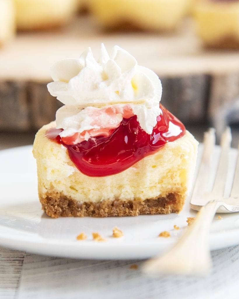 A mini cheesecake cut in half with cherry filling dripping down over the top and whipped cream on top of that.