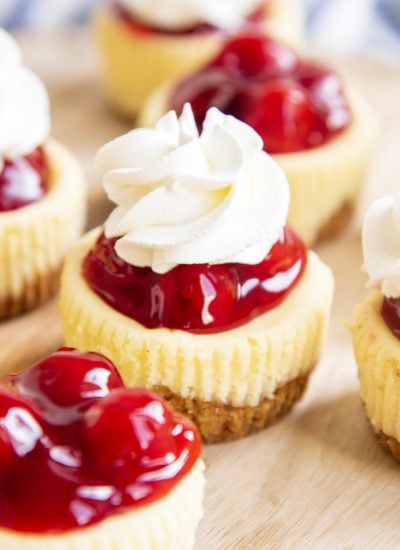 Mini cherry cheesecakes topped with red cherry pie filling on a wooden plate. The cheesecake in the middle has a flower of whipped cream on top.