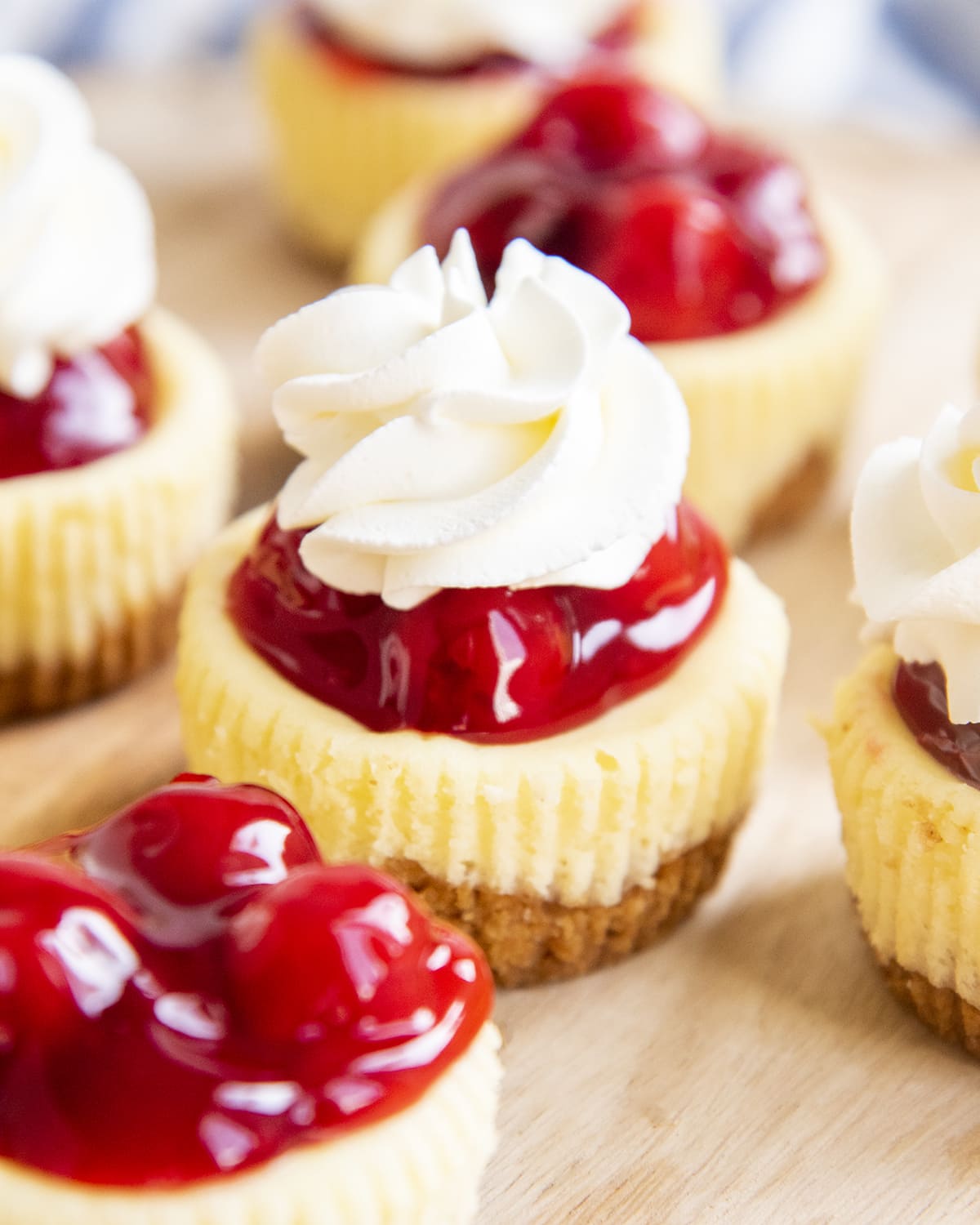 Mini cherry cheesecakes topped with red cherry pie filling on a wooden plate. The cheesecake in the middle has a flower of whipped cream on top.