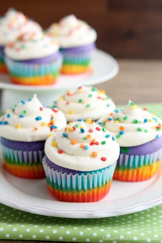Four rainbow cupcakes with white cloud frosting on a plate.