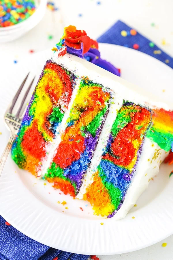A slice of rainbow swirled cake with 3 layers, covered in a white frosting. 