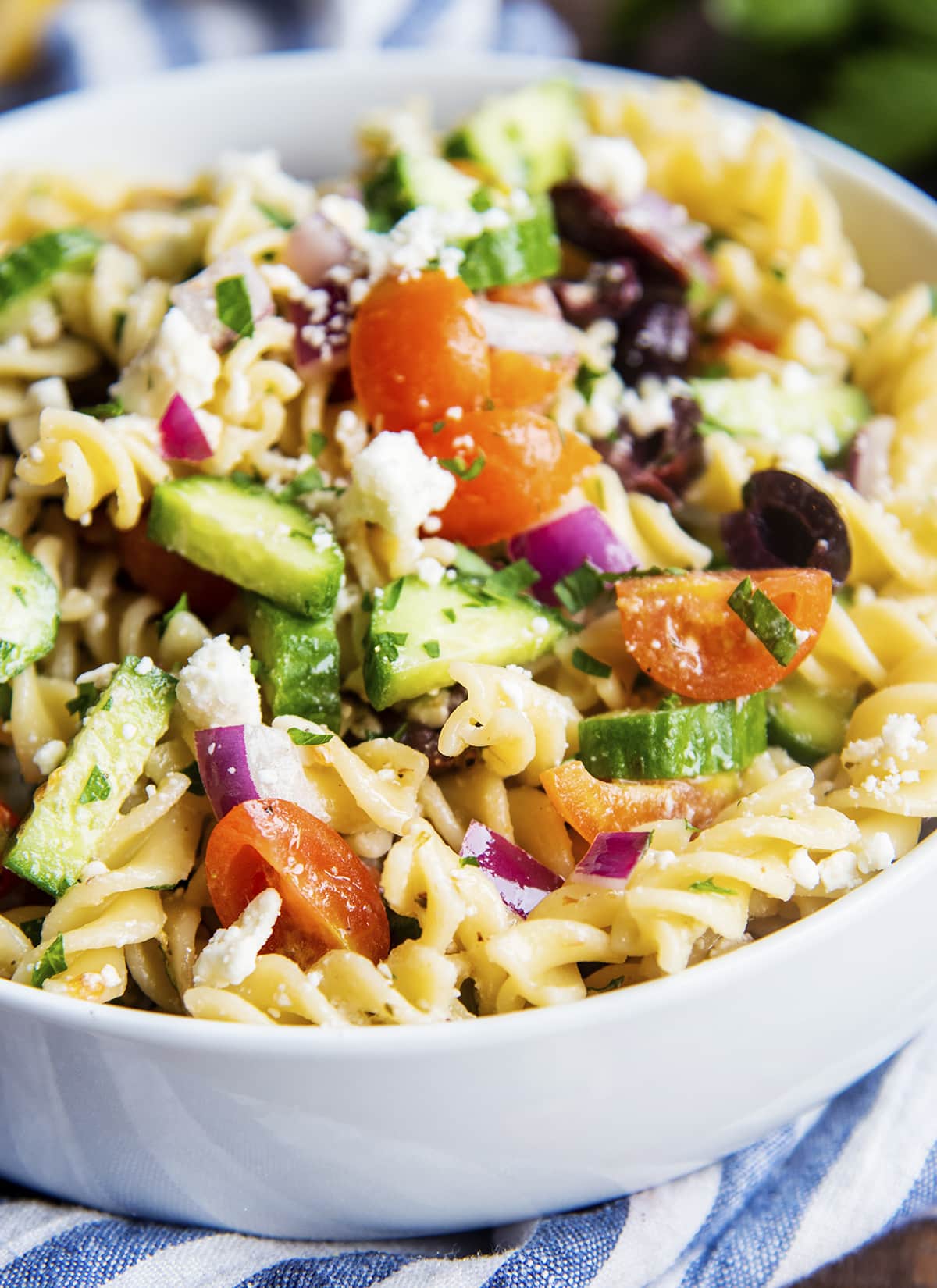 A close up of greek pasta salad with tomatoes, cucumbers, and feta, in a white bowl.