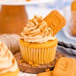 A Biscoff Cupcake on a small wooden stand, with half a Biscoff cookie sticking out of the frositng.