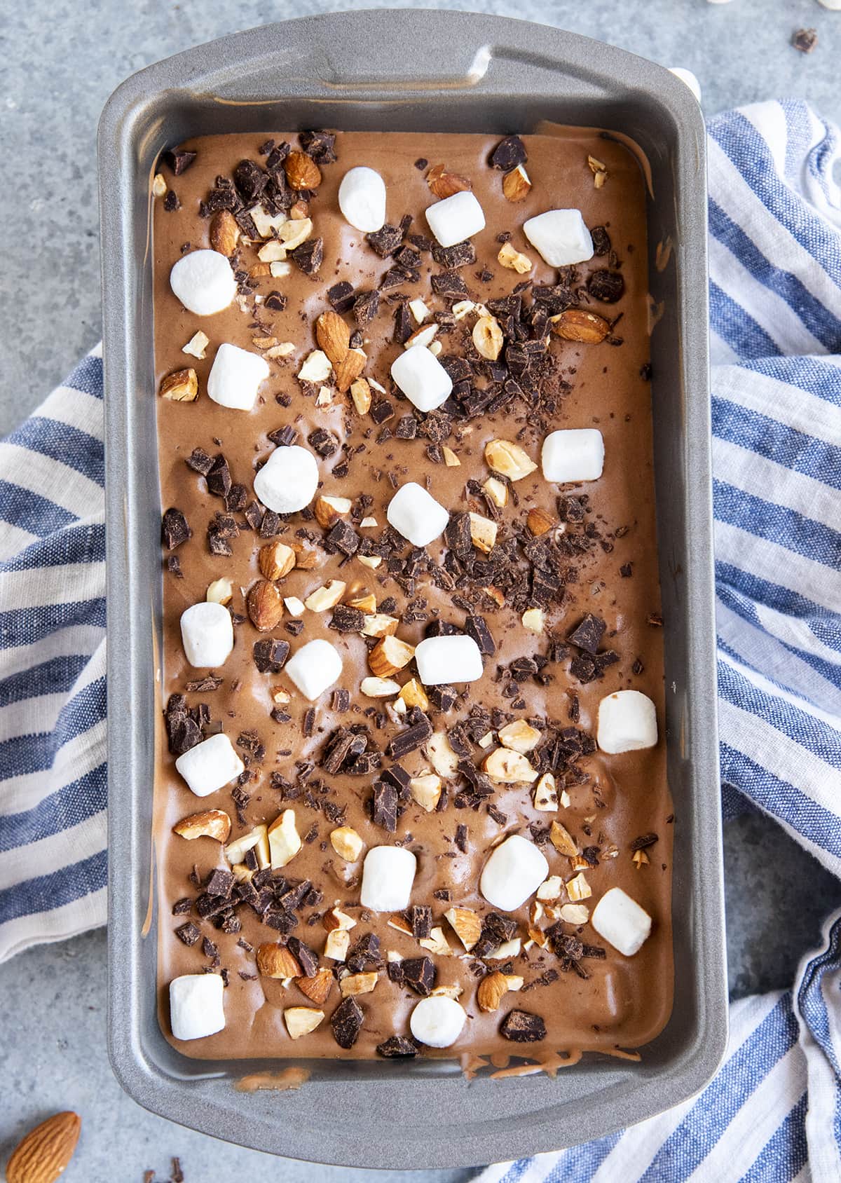 An overhead photo of a metal container full of chocolate ice cream topped with chocolate pieces, mini marshmallows, and chopped almonds.