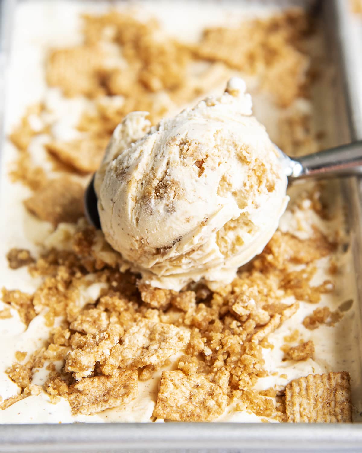 A scoop of cinnamon toast crunch ice cream in an ice cream scoop on top of a container of ice cream. 