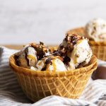 A waffle cone bowl full of scoops of turtle ice cream topped with hot fudge and chopped pecan pieces.