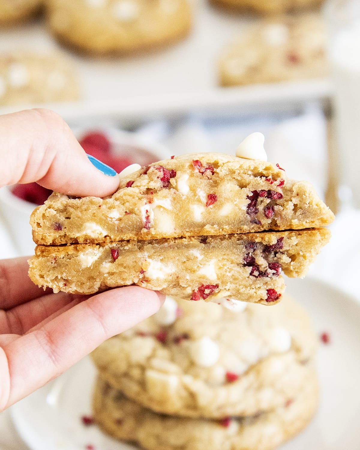A hand holding a giant cookie that is cut in half showing the inside of the cookie. It looks slightly gooey, and there are white chocolate chips and pieces of dried raspberries..