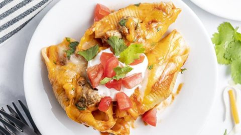 A plate of three Mexican stuffed shells topped with sour cream and diced tomatoes.