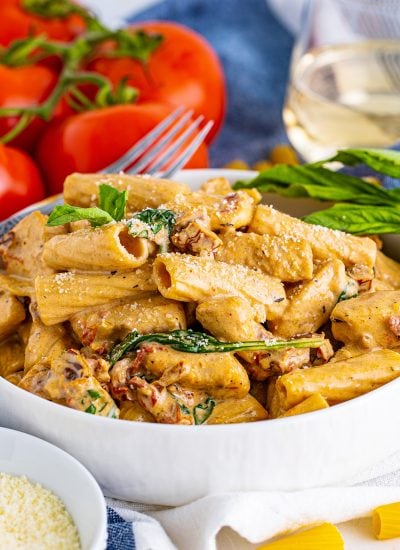 A bowl of chicken rigatoni pasta in a creamy tuscan sundried tomato sauce with tomatoes behind the bowl.