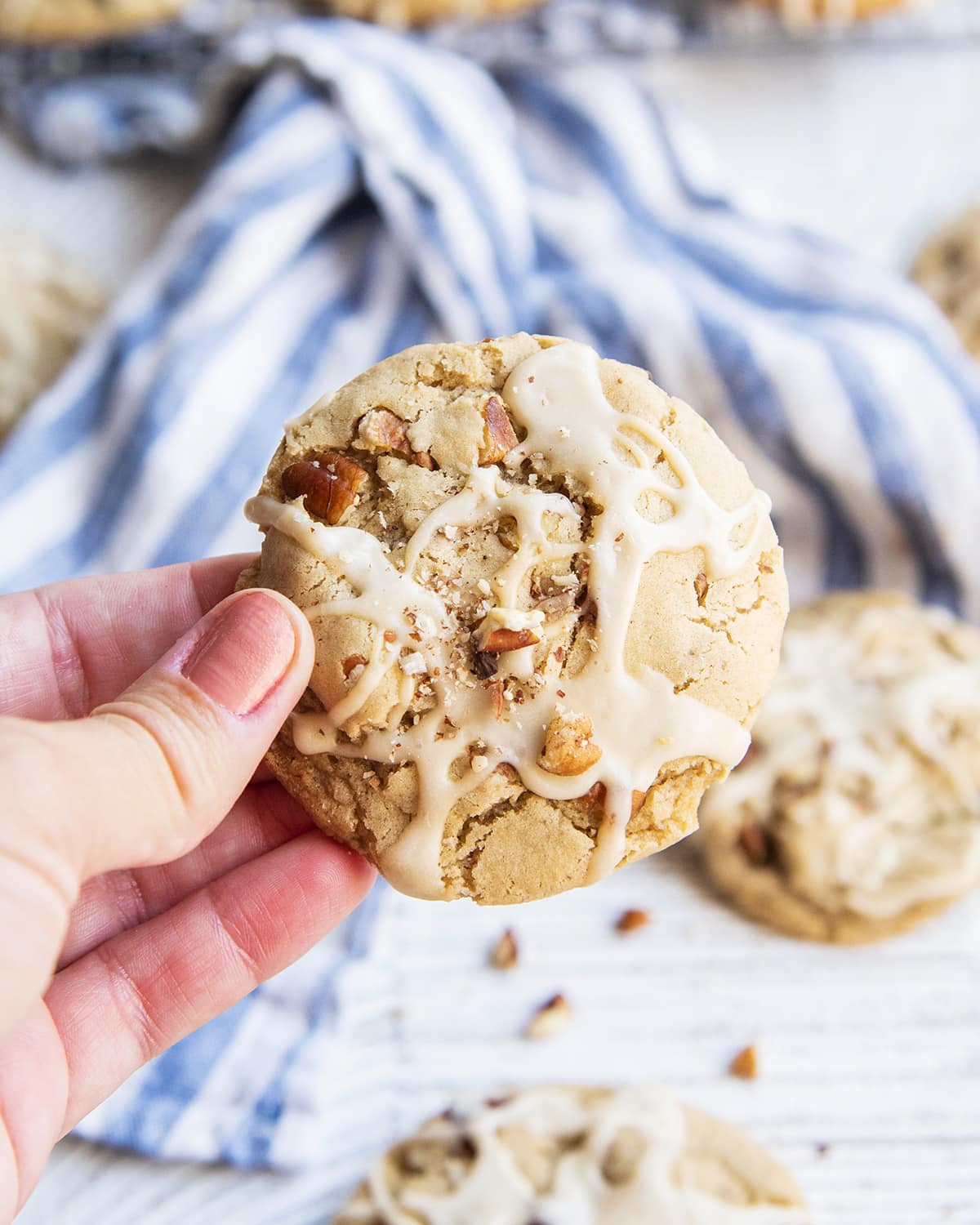 A hand holding a maple pecan cookie topped with a drizzle of maple icing, and pecan crumbs.