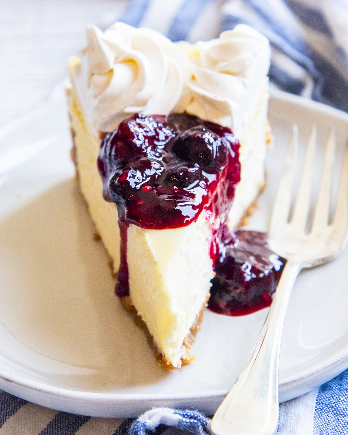 A slice of cheesecake on a plate topped with a berry sauce and whipped cream rosettes.