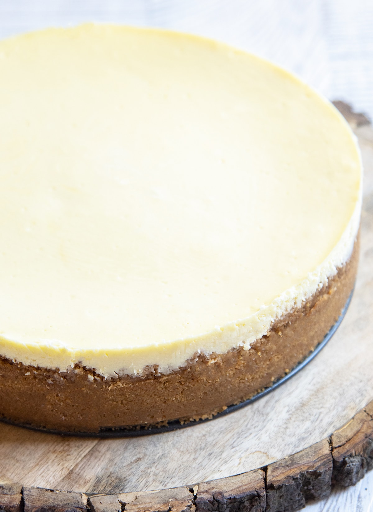 A whole classic cheesecake on a wooden board, with a smooth top, and graham cracker crust on the sides.