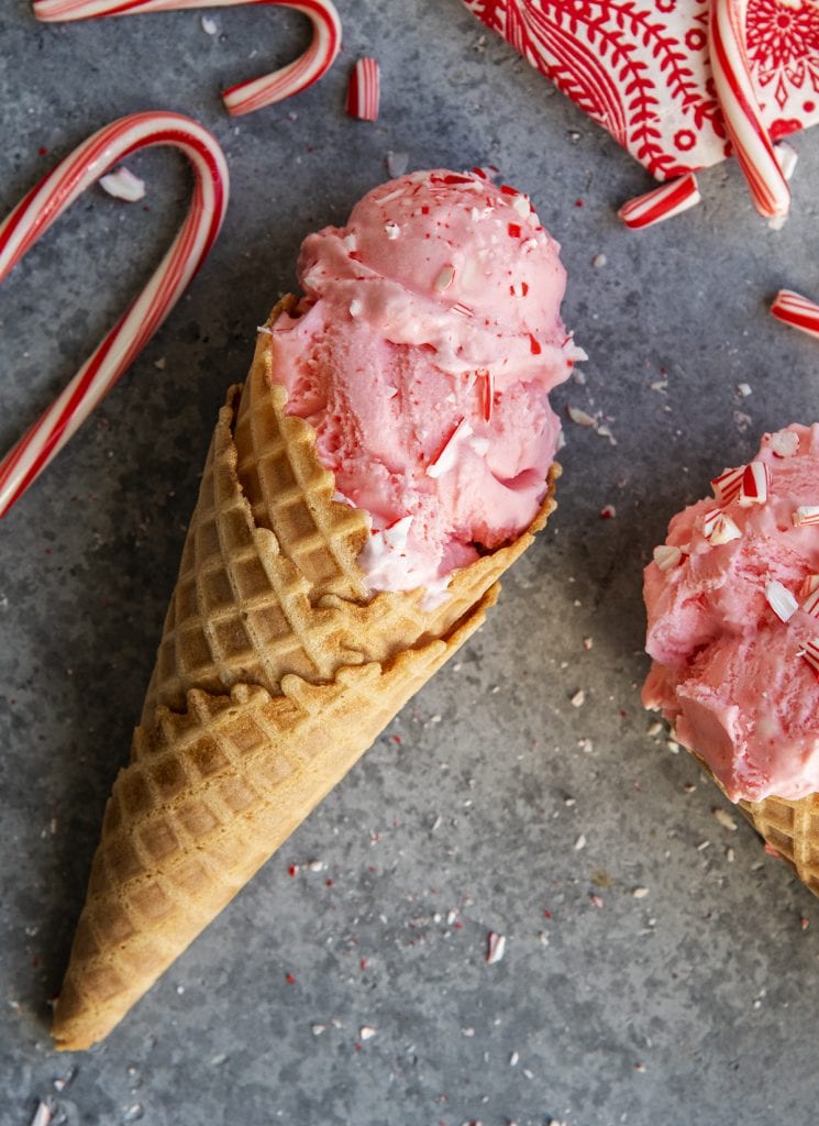 A waffle cone topped with a pink ice cream with candy cane pieces on top laying on a gray counter.