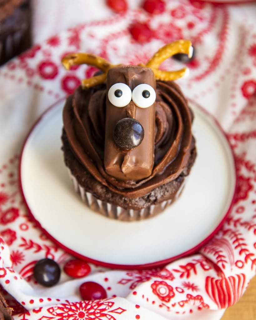 A chocolate cupcake topped with a reindeer with googly eyes and pretzel antlers, and a brown m&m nose.