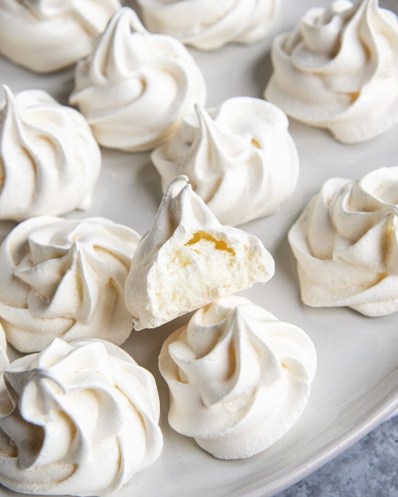 A plate of meringue cookies with one that is bitten in half showing the middle.