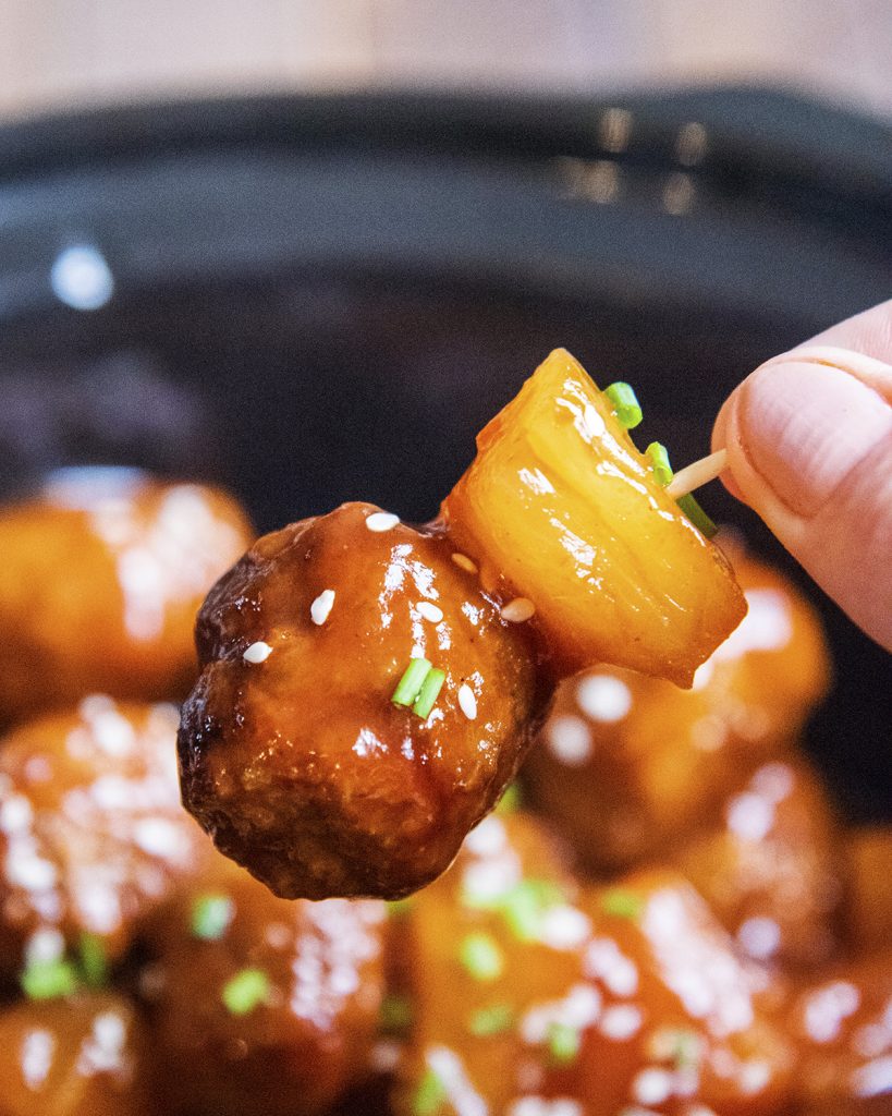 A hand holding a toothpick with a meatball and pineapple on it.