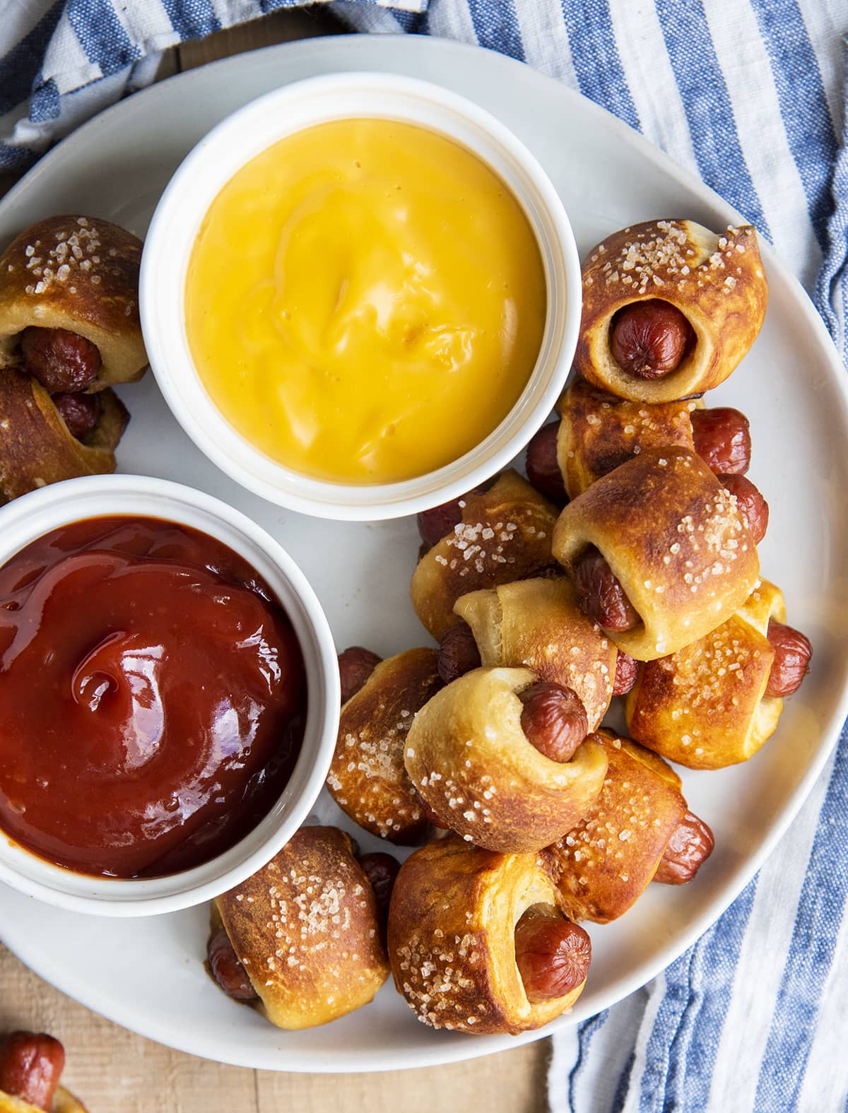 An overhead photo of pretzel pigs in a blanket on a plate with a bowl of ketchup, and a bowl of cheese sauce.