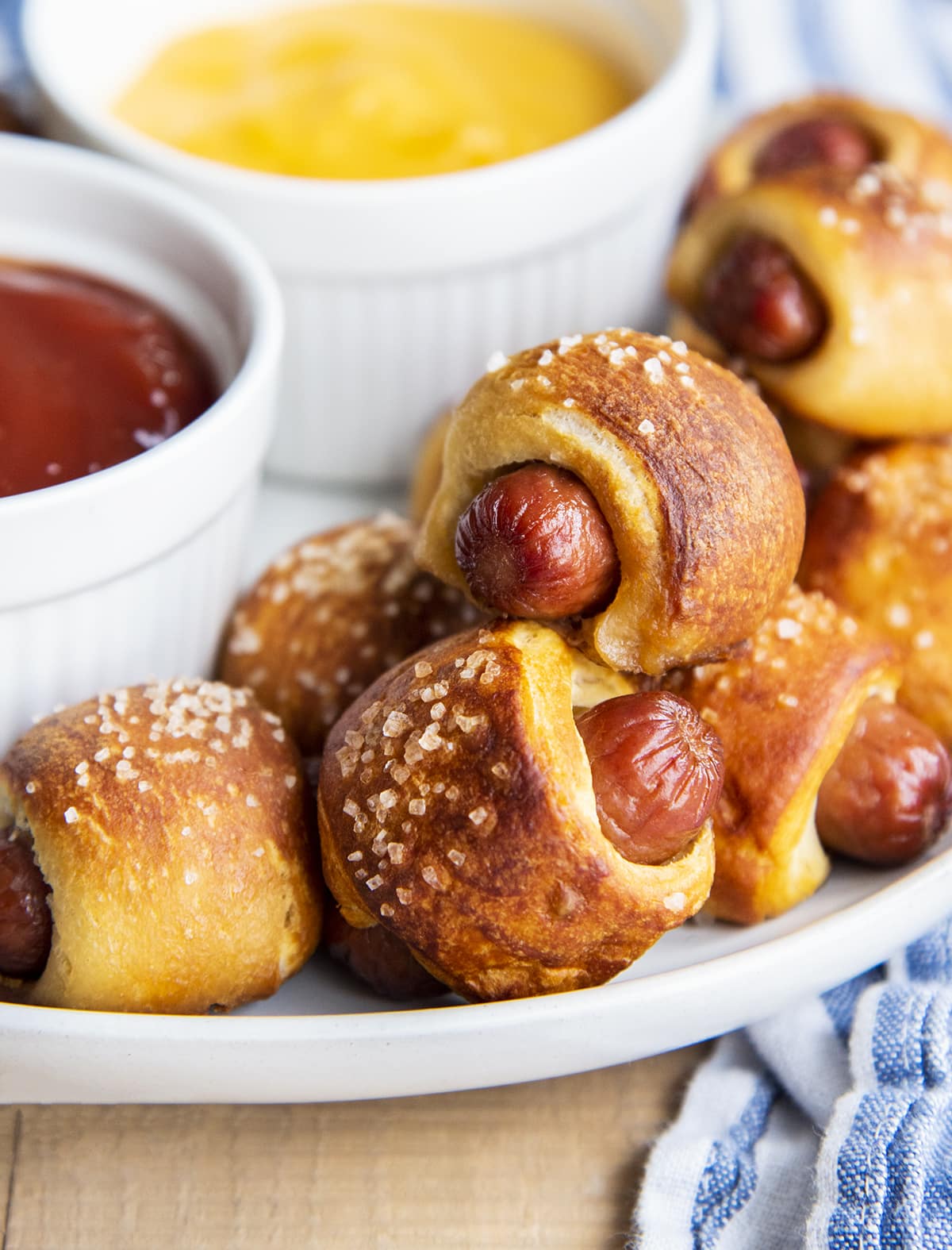 Pretzel pigs in a blanket on a plate with dipping sauces next to them.