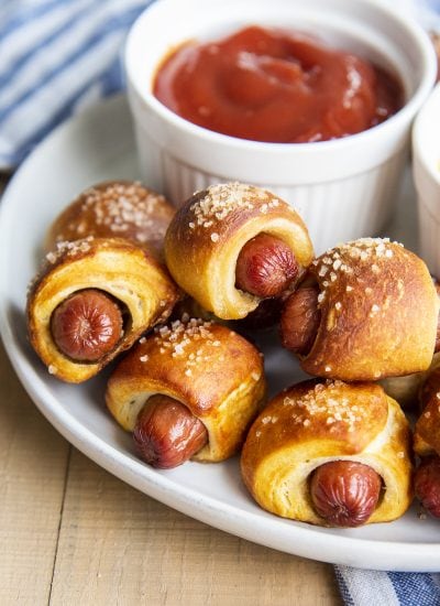 Pretzel pigs in a blanket topped with coarse sea salt on a plate with a bowl of ketchup.