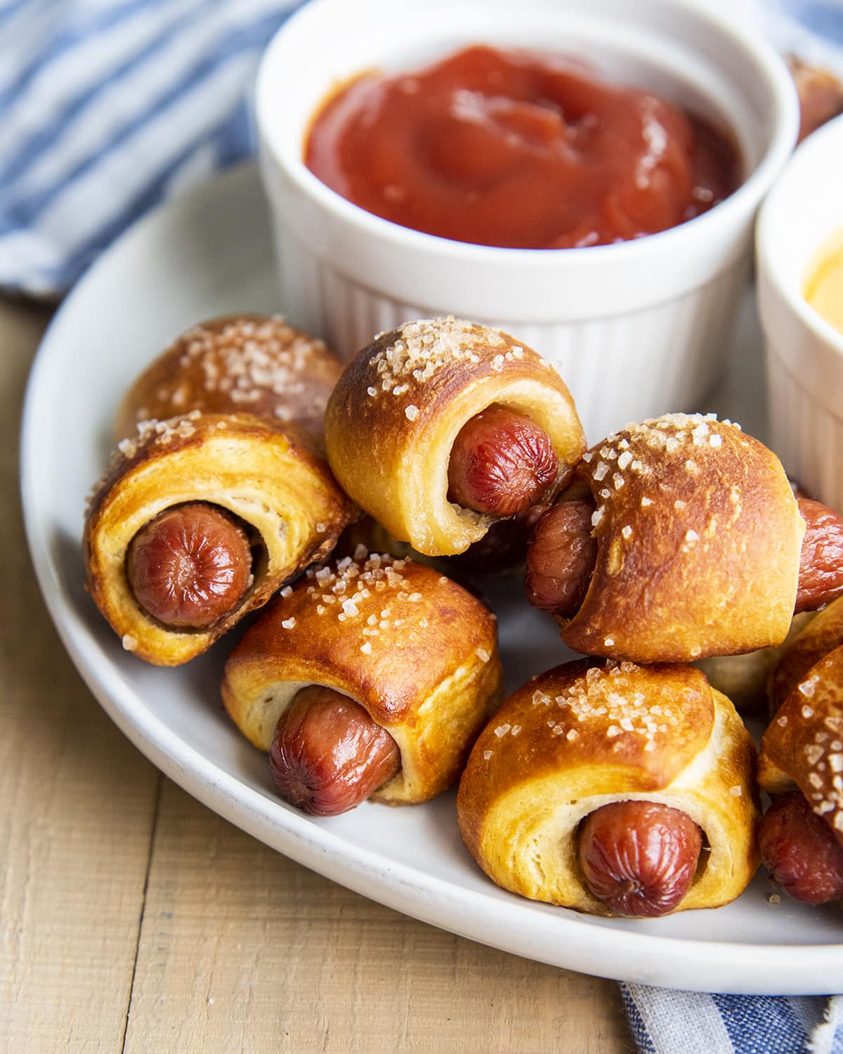 Pretzel pigs in a blanket topped with coarse sea salt on a plate with a bowl of ketchup. 