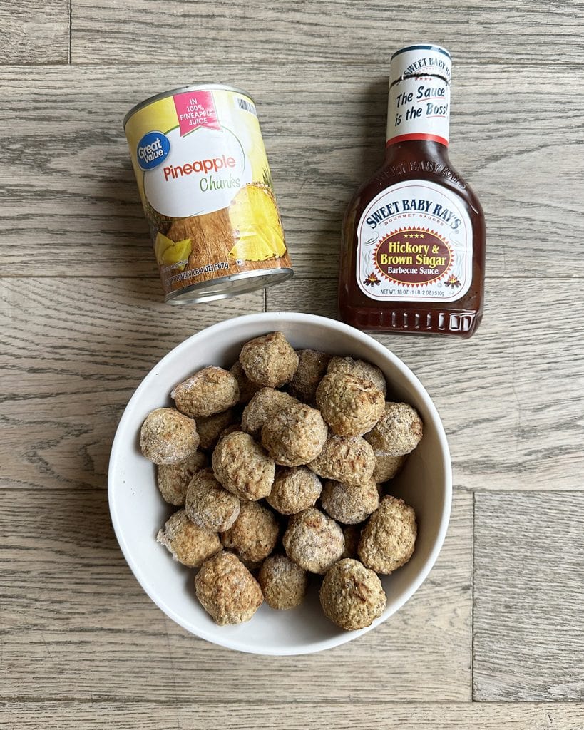 An overhead photo of a can of pineapple chunks, a bottle of bbq sauce, and a bowl of frozen meatballs.