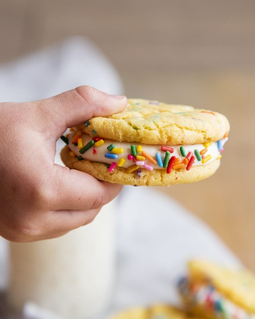 A small hand holding a funfetti sandwich cookie with rainbow sprinkles.