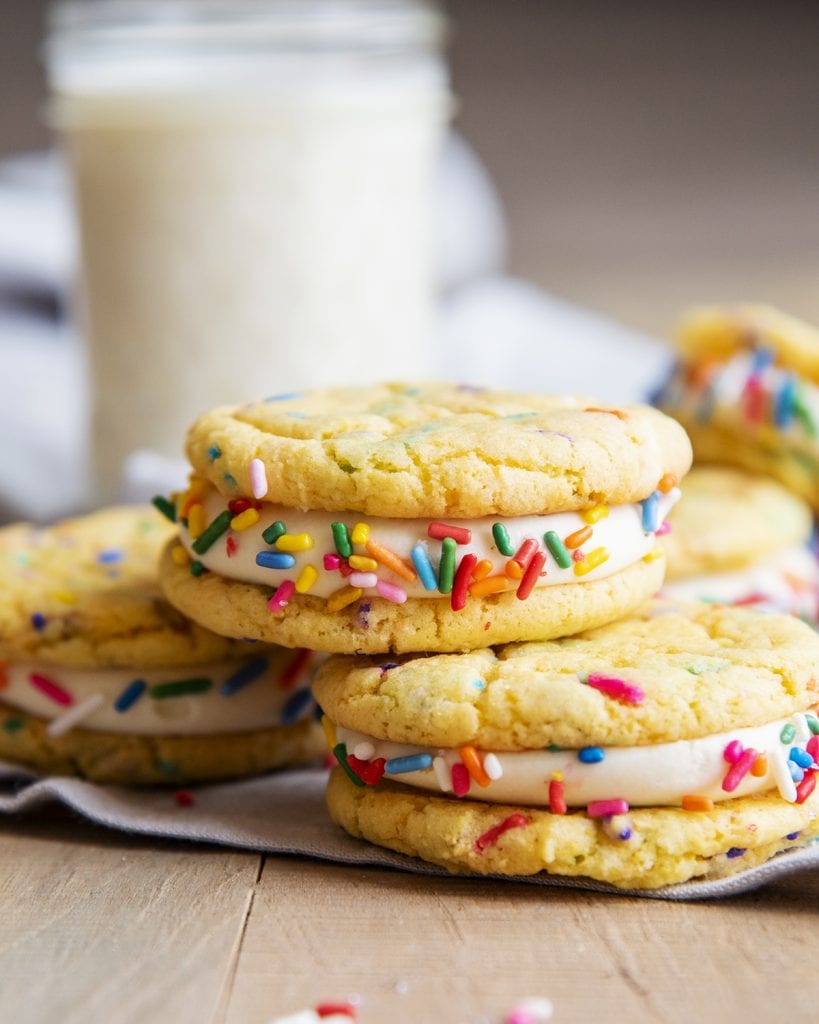 A pile of confetti sandwich cookies with a cup of milk behind them.