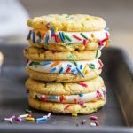 A stack of three cake mix funfetti sandwich cookies with vanilla cream cheese frosting and rainbow sprinkles on a cookie sheet.