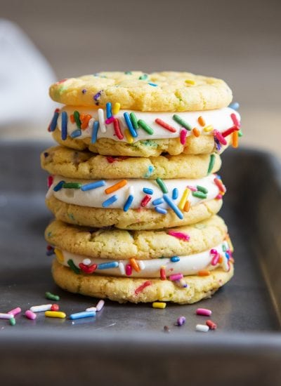 A stack of three cake mix funfetti sandwich cookies with vanilla cream cheese frosting and rainbow sprinkles on a cookie sheet.