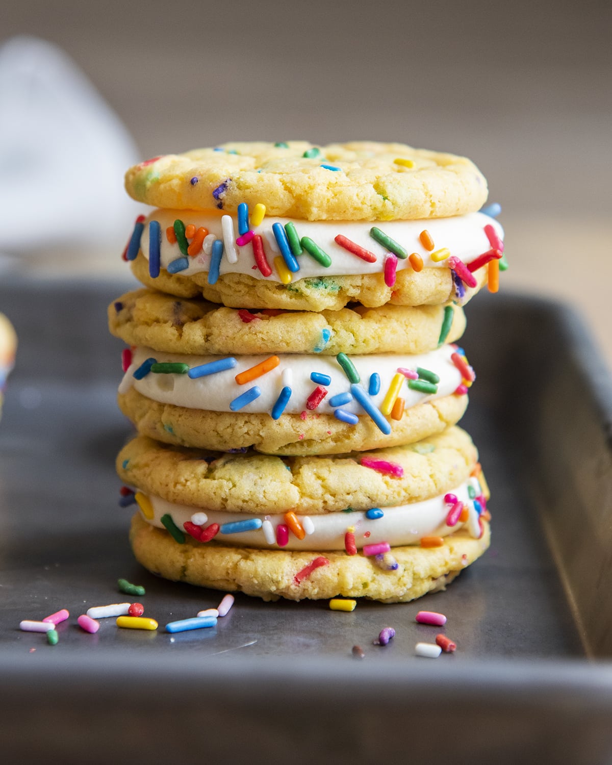 A stack of three cake mix funfetti sandwich cookies with vanilla cream cheese frosting and rainbow sprinkles on a cookie sheet.