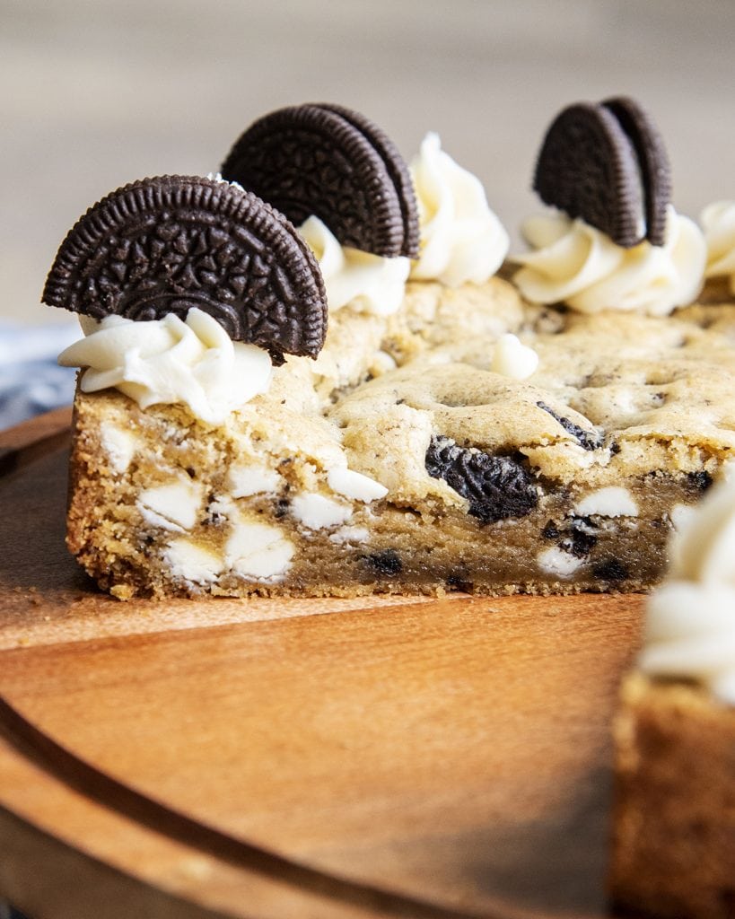 A cut open cookie cake on a wooden board. It is filled with white chocolate chips and Oreo pieces.