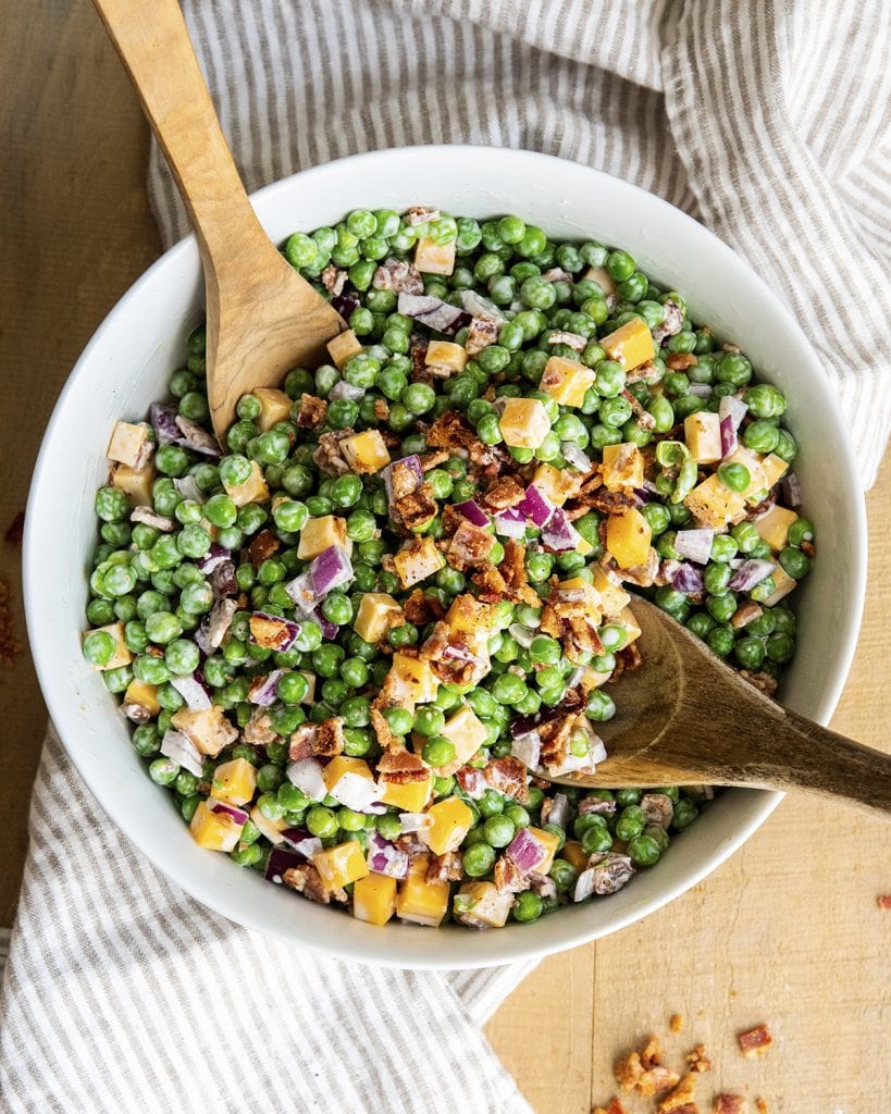 An overhead photo of a bowl of creamy bacon pea salad with two wooden spoons in the bowl.