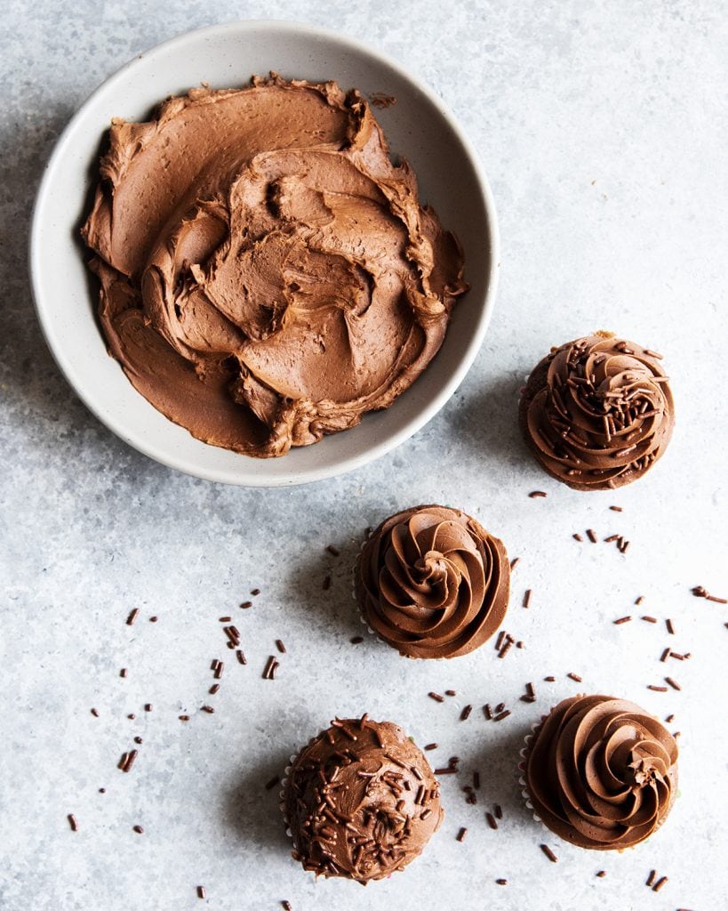 A bowl of chocolate frosting next to 4 chocolate frosted chocolate cupcakes.