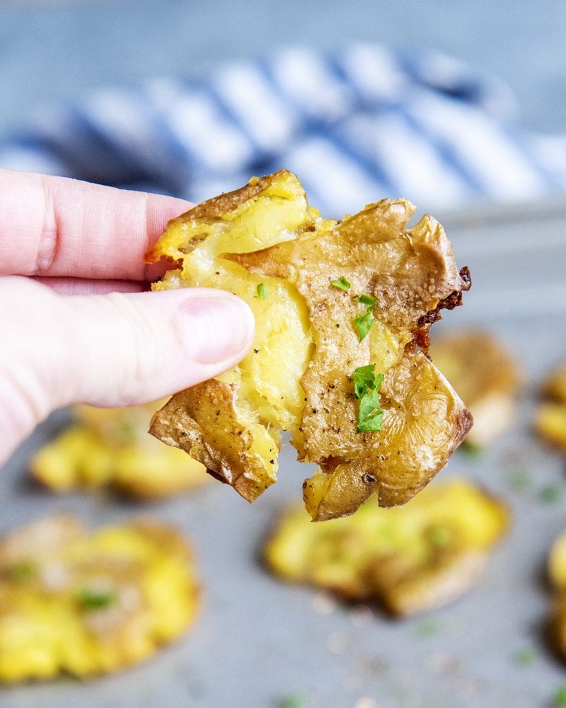 A hand holding a crispy smashed potato topped with fresh parsley.