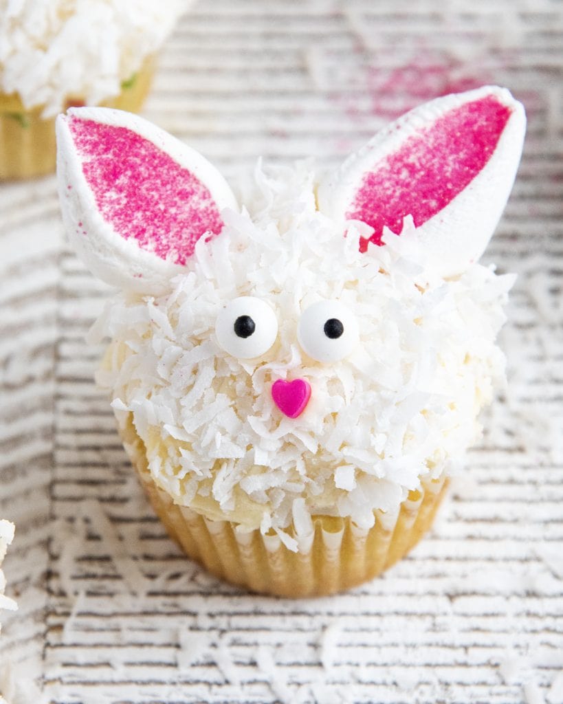 A cupcake decorated to look like an Easter bunny with marshmallow ears, candy eyes, and a sprinkle nose.
