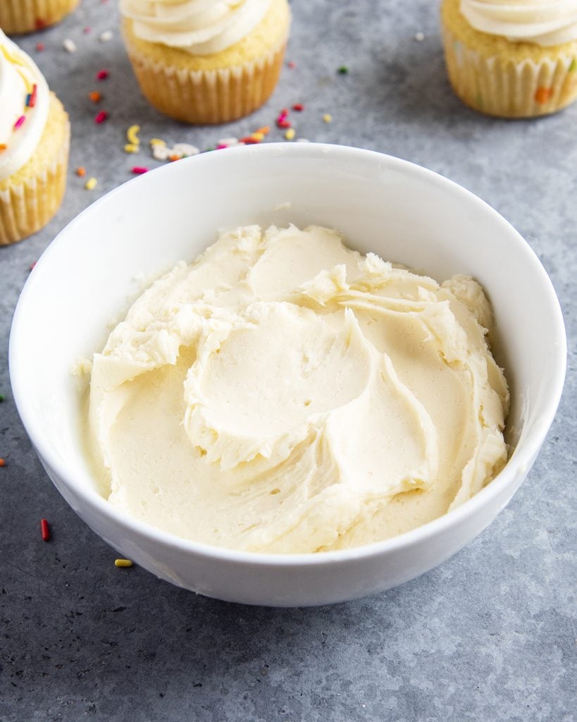A bowl of vanilla buttercream frosting, with some frosted cupcakes behind it.