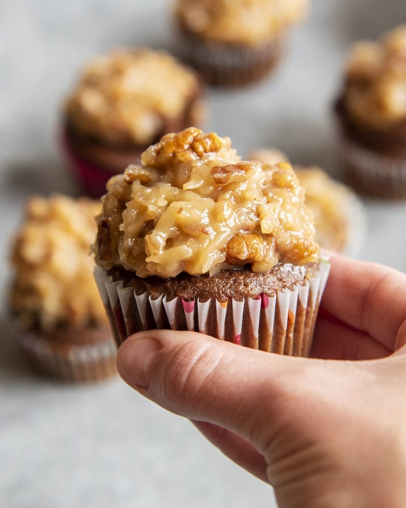 A hand holding a chocolate cupcake topped with coconut pecan frosting.