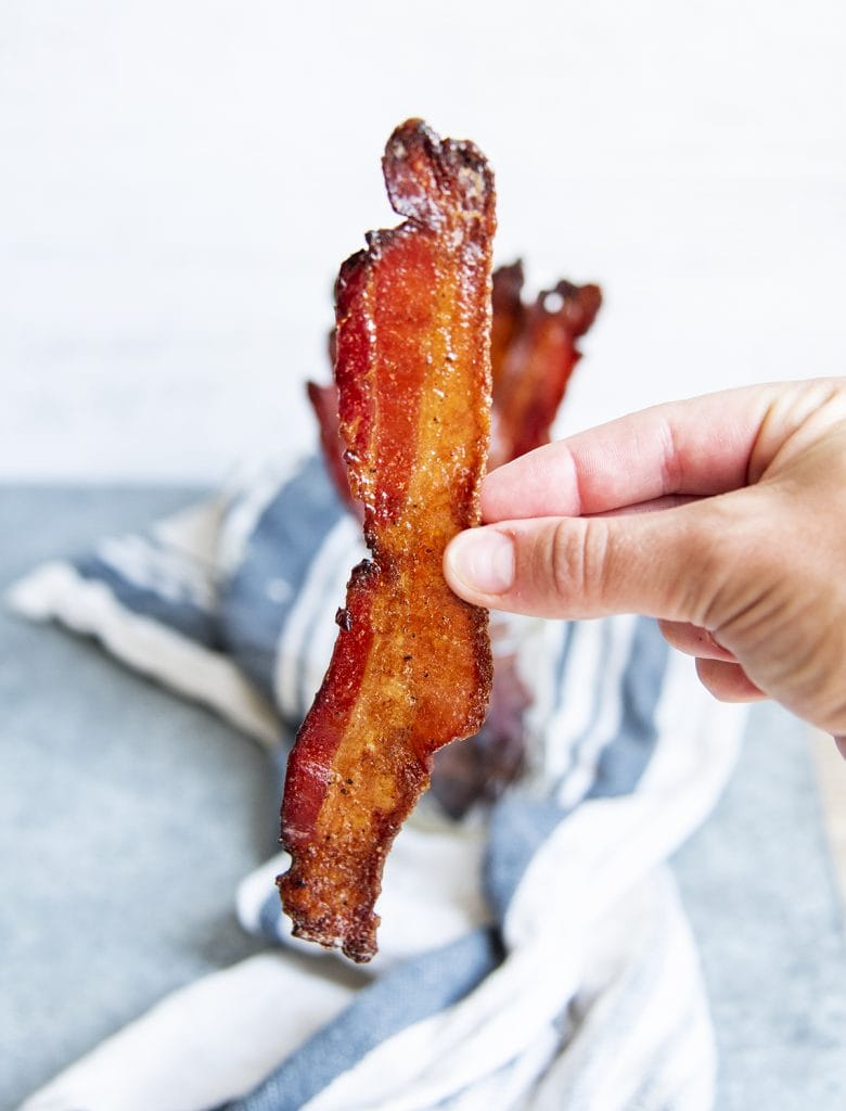 A hand holding a strip of brown sugar candied bacon.