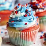 A close up of a cupcake with red, white, and blue cake batter, and swirled frosting on top with patriotic sprinkles on top.