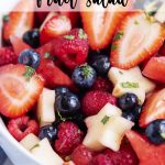 A fruit salad with red, white, and blue fruit in a white bowl, topped with fresh mint pieces, with a text overlay for pinterest.