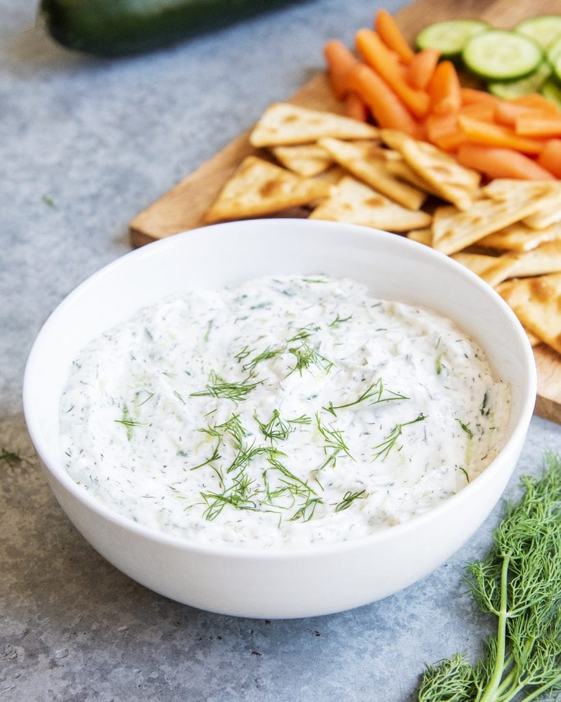 A bowl of homemade tzatziki with a tray of chips, and vegetables behind it.