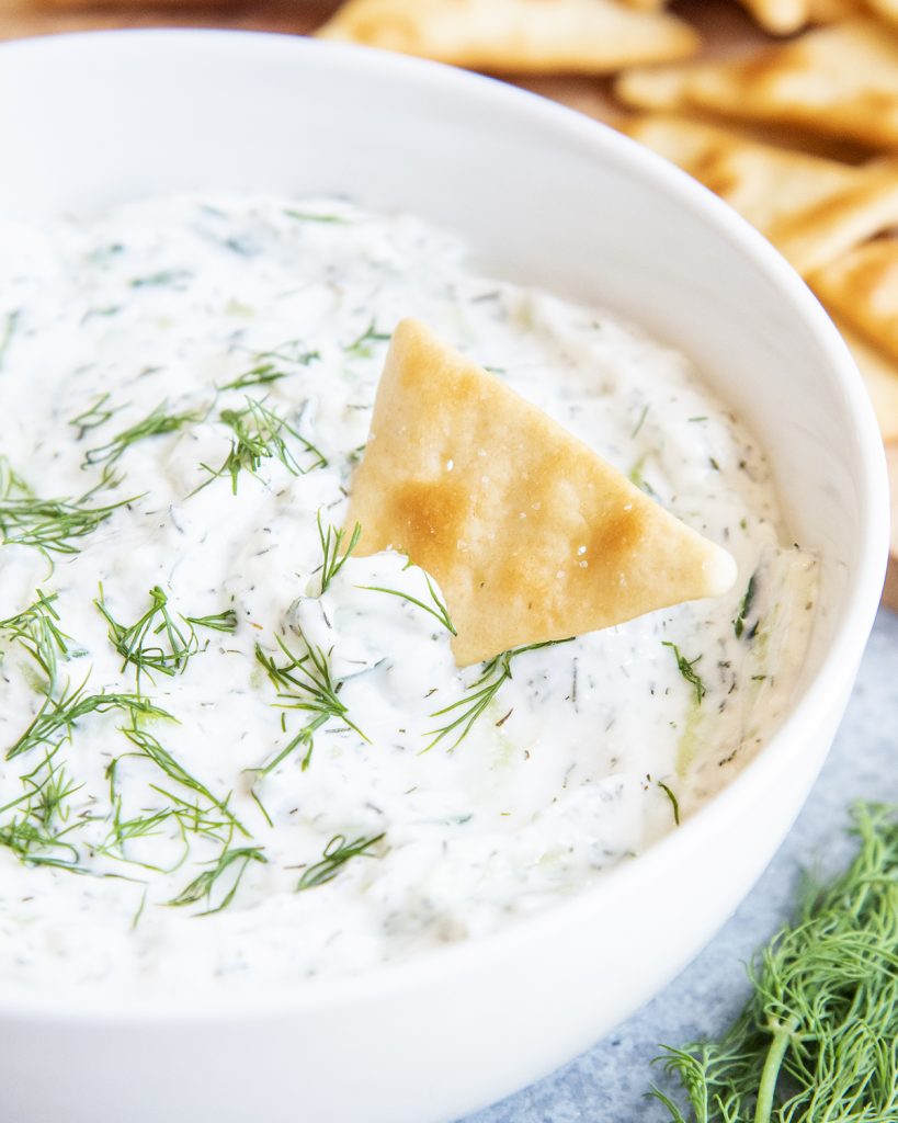A close up of a bowl of tzatziki sauce topped with fresh dill, and with a pita chip in the bowl.
