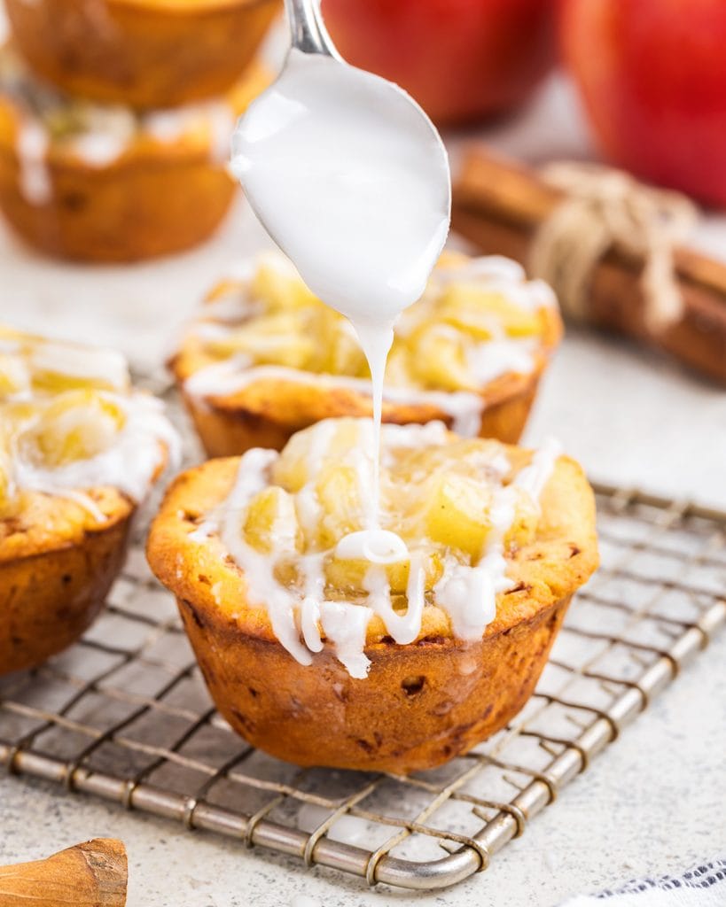 Vanilla icing being drizzled by a spoon onto a cinnamon roll apple pie cup.