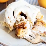 A peach cinnamon roll on a plate, topped with cream cheese frosting, and with a bite cut out of it on a fork.