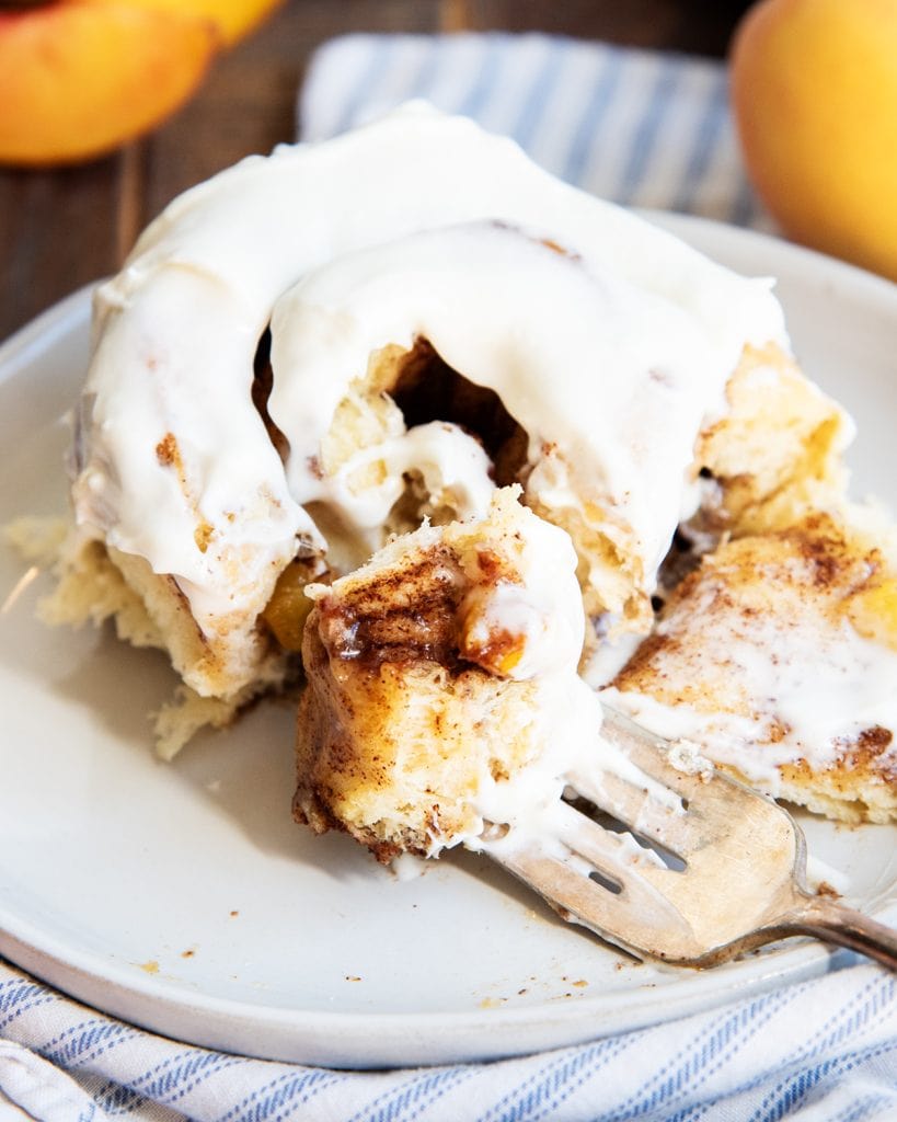 A peach cinnamon roll on a plate, topped with cream cheese frosting, and with a bite cut out of it on a fork.