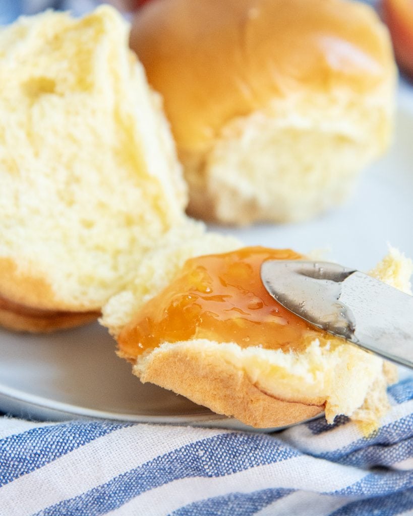A roll being slathered with peach jam with a knife. 