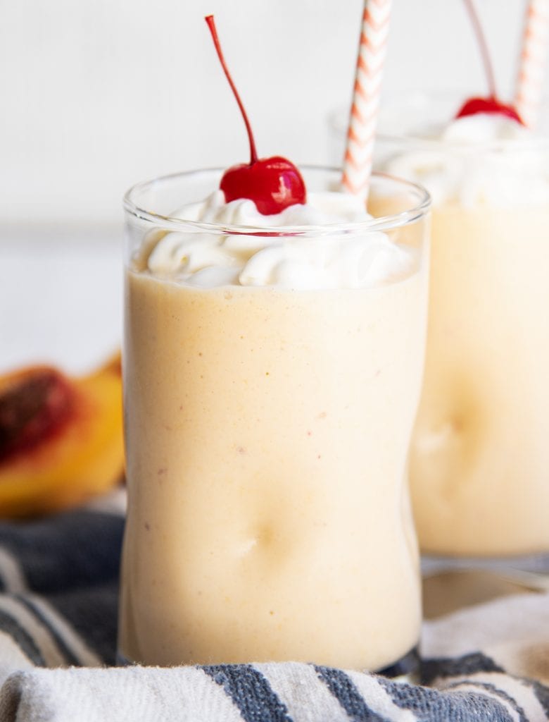 A close up of a glass full of peach milkshake, and topped with whipped cream and a maraschino cherry.